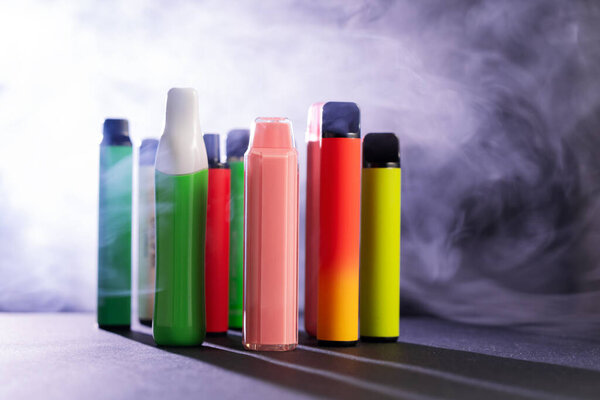 A set of colorful disposable electronic cigarettes of different shapes on a black background with smoke. Concept of modern smoking, vaping and nicotine..