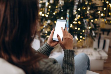 Mesaj atmak için yeri olan bir Noel modeli. Noel reklamı, uygulama şablonu. Telefonun arkasında boş bir ekran olan el ele tutuşmuş altın gibi güzel bir Noel ağacının akşam ışıkları altında.. 