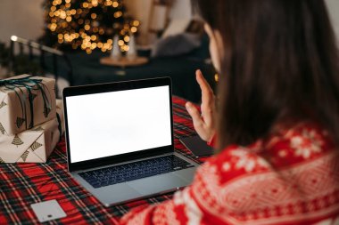 Şenlikli dekorasyonlar, hediyeler ve Noel ışıklarıyla bir video görüşmesi sırasında dizüstü bilgisayara el sallayan bir kadın, rahat bir tatil ortamı yaratıyor.. 