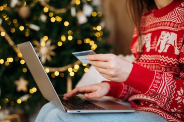 Şenlikli süveter dükkanlarında kredi kartı ve dizüstü bilgisayarı olan bir kadın. Arka planda Noel ağacı ışıkları var.. . 