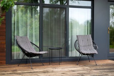 İki modern sandalye ve cam kapıların yanındaki ahşap terasta küçük bir masa. Rahatlamak için açık hava oturma alanı.