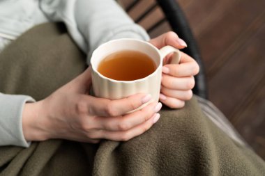 Bir kadın battaniyenin altında sıcak bir fincan çay tutarken, serin bir günde elini ısıtıyor. 