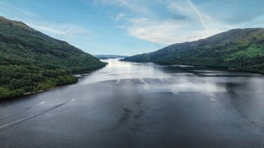 Lomond Gölü, İskoç dağları, berrak mavi gökyüzünün altında tüylü beyaz bulutlarla sakin bir göl manzarası. Yoğun ağaçlarla bezenmiş yemyeşil tepeler suları çevreleyerek, huzurlu bir manzara yaratıyor.