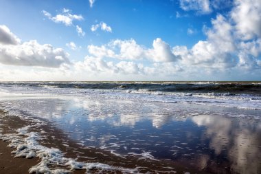 Bu sakin sahil sahnesinde engin bir kumlu sahil manzarası var dramatik mavi gökyüzünün yansımaları ve dağınık bulutlar, yumuşak okyanus dalgalarıyla buluşuyor..