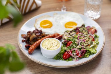 Kahvaltı seti, bir restoranda. Kızarmış yumurta, sosis, sosis, kızarmış pastırma, sos ve salata, bir tabakta, seçici bir odak noktası..