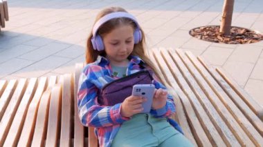Sarışın çocuk kulaklıkları. Damalı gömlek. Şehir merkezinin dışında müzik podcast akıllı telefon. Konsantre kız çevrimiçi sesli medya 4K. Ruh sağlığı başvurusu Stres destek uygulaması