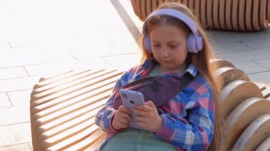 Sarışın çocuk kulaklıkları. Damalı gömlek. Şehir merkezinin dışında müzik podcast akıllı telefon. Konsantre kız çevrimiçi sesli medya 4K. Ruh sağlığı başvurusu Stres destek uygulaması