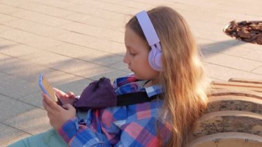 Sarışın çocuk kulaklıkları. Damalı gömlek. Şehir merkezinin dışında müzik podcast akıllı telefon. Konsantre kız çevrimiçi sesli medya 4K. Ruh sağlığı başvurusu Stres destek uygulaması