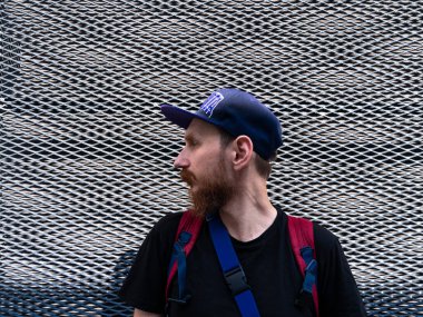 Metal çitin arka planında sakallı, bin yıllık hippi bir adamın profil portresi. Şık, kendine güveni olan, kızıl sakallı, sıradan kıyafetleri olan, güneşli şehir caddesinde yürüyen bir adam..