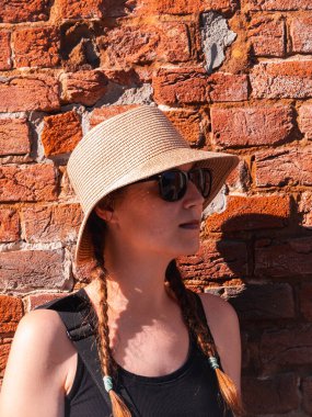 Kırmızı tuğlalı duvar kentsel arka planında hasır şapkalı genç bir kadın. Milenyum kızı güneş ışığıyla Summer City caddesinde yürüyor. Siyah güneş gözlüklü, kendine güvenen, şık bir bayan..