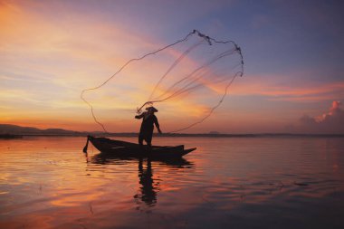 Balıkçı gün batımında ağlarını örüyor. Siluet Asyalı balıkçı ahşap teknede tatlı su balıkları için ağ atıyor. Tayland kültürü.