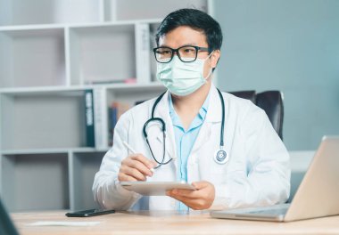 Tıbbi maske takan mutlu erkek doktorun portresi kendi muayenehanesinde, beyaz önlüklü doktor veya kardiyolog ofisinde masasında oturuyor.