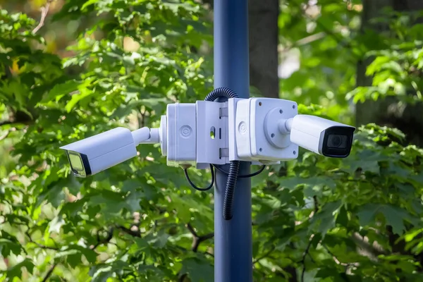 Two outdoor city video security cameras on a pole on background of green leaves. Safe and comfortable urban environment. Control and combating crime. Street security, safety and city life improvement.