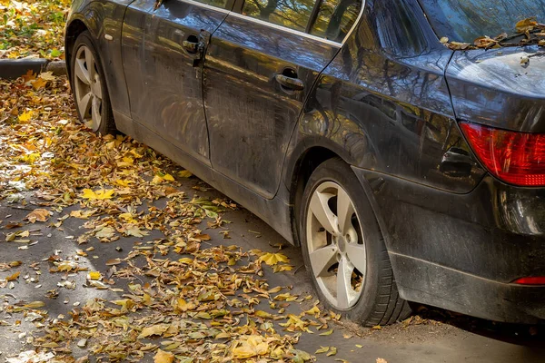 An abandoned dusty car with flat or tear tires in a city parking and yellow autumn leaves. The broken rust vehicle in the courtyard of an apartment building. Unecological unauthorized car dump.