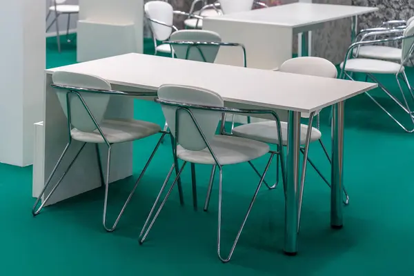 White office tables and chairs with chrome legs on green carpet. A comfortable environment for communication between participants of corporate events, exhibitions, conferences or company employees.