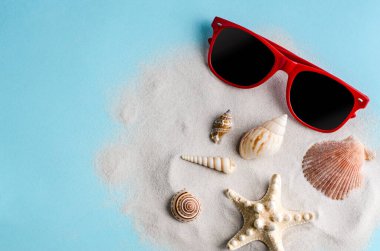Mavi arka planda temiz kum üzerinde güneş gözlüğü ve deniz kabukları. Mevsimlik, yaz tatili ve eğlence konsepti. Düz döşeme, kopyalama alanı, metin için yer. Yukarıdan görüntüle.