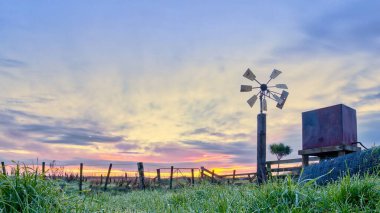 Çiftlik rüzgar pompası yanında su kulesiyle biraz yıpranmış görünüyor. Arka planda harika bir gündoğumu gökyüzü var..