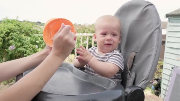 女人用勺子喂累了的孩子 妈妈给哭哭啼啼的可怜的小男孩提供纯芦苇食物 妈妈在花园里的婴儿椅上喂孩子 夏天的时候幼儿营养 — 图库视频影像