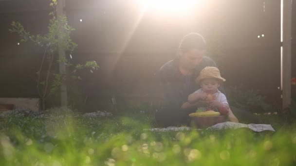 緑の草で果物を食べる赤ちゃんを保持する幸せな若い陽気な母親 裏庭で愛を持って屋外で遊んでママ愛らしい幼児 両親を持つ小さな子供 生態学的概念 — ストック動画