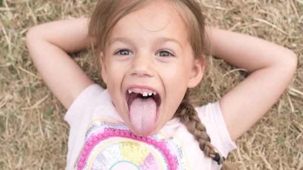 Little Happy Toothless Smiling Child Girl Laying on Yellow Lawn Dry Grass Hay in Park Shows Tongue. Summer Time, Nature, Lifestyle Country life farm village. Portrait Face Close up Looking at Camera.