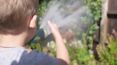 Bahçenin arka bahçesindeki çim bitkilerini sulayan komik küçük çocuk. Sıcak güneşli bir yaz gününde sulama hortumuyla oynayan sevimli bir çocuk. Çocuklar ev işlerine yardım eder. Çocuklar için etkinlik. Çocukluk.