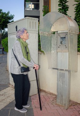 Gri ve yeşil giyinmiş yaşlı bir kadın sökülmüş bir ankesörlü telefonun yanında baston ile duruyor. Nostaljik bakışları kaldırım ve yeşillik manzarasıyla çelişiyor..