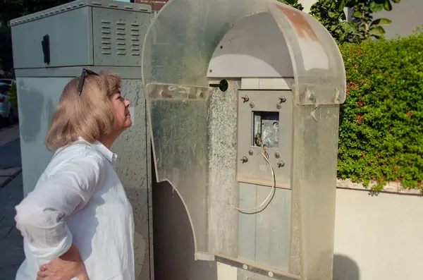 stock image Wearing sunglasses on her head, a woman leans in to inspect a dilapidated payphone, now just a metal and plastic shell with exposed wires. The urban backdrop includes greenery and electrical boxes.