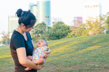 Asyalı anne, bebeğini şehir parkında gün batımı aktivitesinde tutuyor.