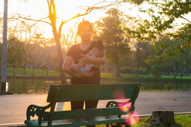 Asyalı anne, bebeğini şehir parkında gün batımı aktivitesinde tutuyor.
