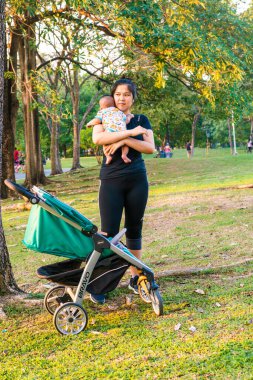 Asyalı anne, bebeğini şehir parkında gün batımı aktivitesinde tutuyor.