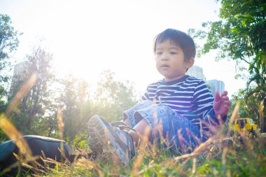 Yeşil çimenlerde oturan mutlu Asyalı çocuk bitkilerin doğasıyla oynuyor.
