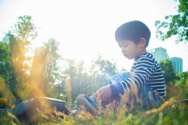 Yeşil çimenlerde oturan mutlu Asyalı çocuk bitkilerin doğasıyla oynuyor.