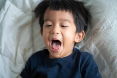 4 yaşındaki Asyalı çocuk beyaz yatakta mutlu bir şekilde yatarken gülüyor.