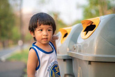 Genç Asyalı çocuk çevreyi temizleyip çöpleri parktaki bir çöp kutusuna atıyor.