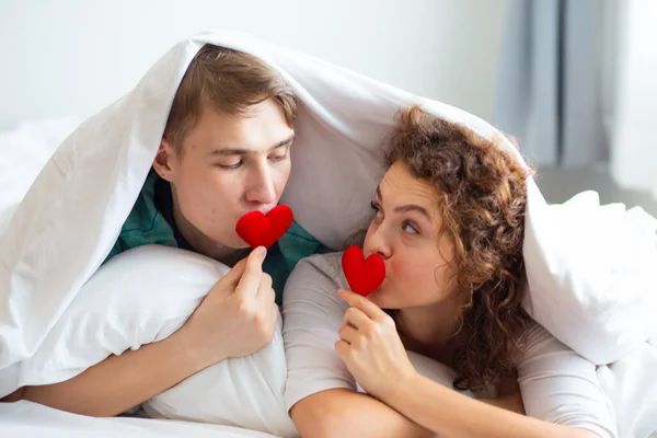 Love couple lying on bed under blanket holding red hearts, happy romantic life