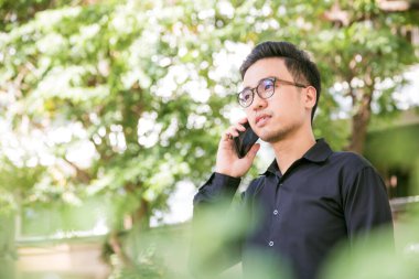 Mutlu genç yakışıklı adam stand altında açık havada ağaç ve iş için smartphone kullanarak konuşmak, gözlük adam