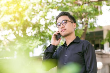 Mutlu genç yakışıklı adam stand altında açık havada ağaç ve iş için smartphone kullanarak konuşmak, gözlük adam