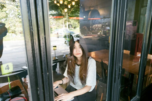 年轻迷人的亚洲妇女坐在镜子窗口和打开笔记本电脑在现代咖啡店 — 图库照片