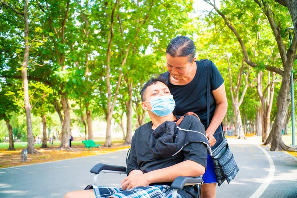 Disabled man sitting in a wheelchair wear mask enjoying in the public park with mom outdoor vacation