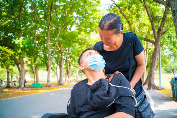 Disabled man sitting in a wheelchair wear mask enjoying in the public park with mom outdoor vacation