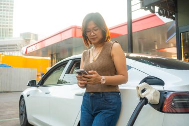 Genç, güzel Asyalı bir kadın elektrik istasyonunda duruyor, kablolu interneti kuruyor, akıllı telefondan internete bağlanıyor ve elektrikli araba ile seyahat ediyor.