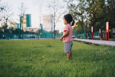 Güzel küçük Asyalı kız şehir parkında gün batımında yeşil çimlerde yürüyüp koşuyor.