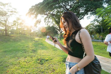 Business casual asian woman use smartphone in city park sunset light online business communication clipart