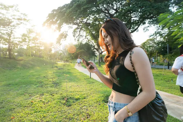 ビジネス カジュアルなアジアの女性は都市公園の日没ライトのオンラインビジネスコミュニケーションのスマートフォンを使用します ストック写真