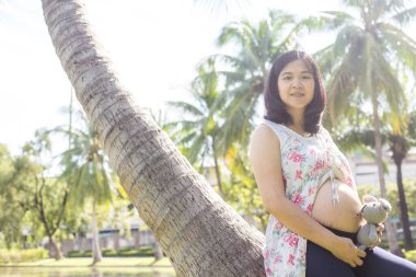 Genç, mutlu, hamile, Asyalı kadınlar parkta oyuncak bebekle rahatlıyor.