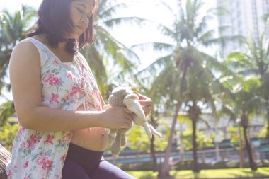 Genç, mutlu, hamile, Asyalı kadınlar parkta oyuncak bebekle rahatlıyor.