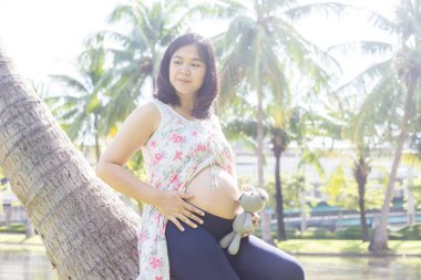 Genç, mutlu, hamile, Asyalı kadınlar parkta oyuncak bebekle rahatlıyor.