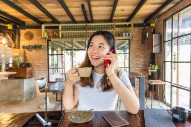 Güzel gülümseyen Asyalı kadın internet üzerinden kahve içerken akıllı telefondan konuşuyor.