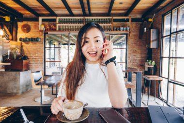 Güzel gülümseyen Asyalı kadın internet üzerinden kahve içerken akıllı telefondan konuşuyor.