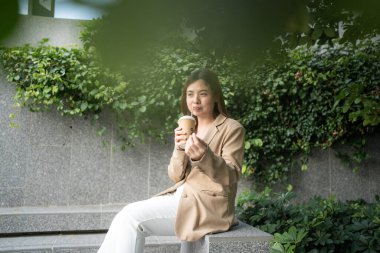 Girişimci Asyalı kadın kahve içiyor. Şehir merkezinin önünde akıllı telefon kullanıyor.